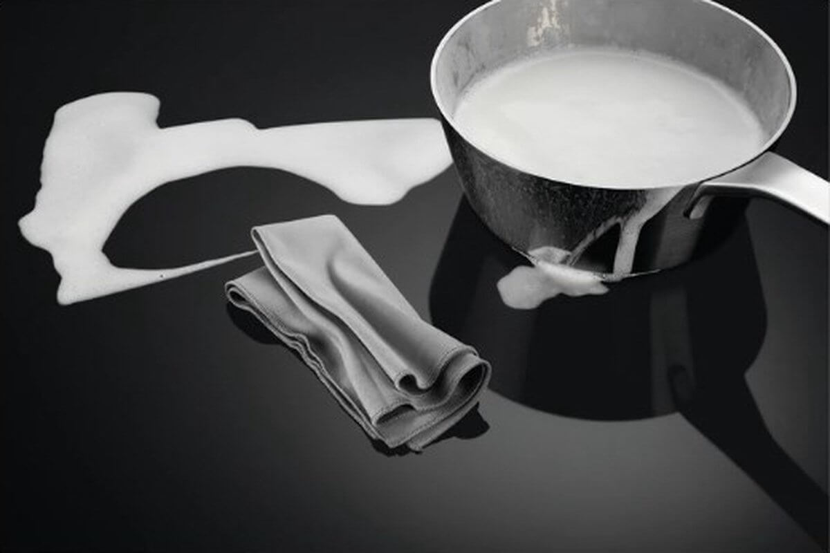 Overgekookte melk op de AEG IKB84443IB inductiekookplaat met schoonmaakdoek, toont eenvoudig schoon te maken oppervlak