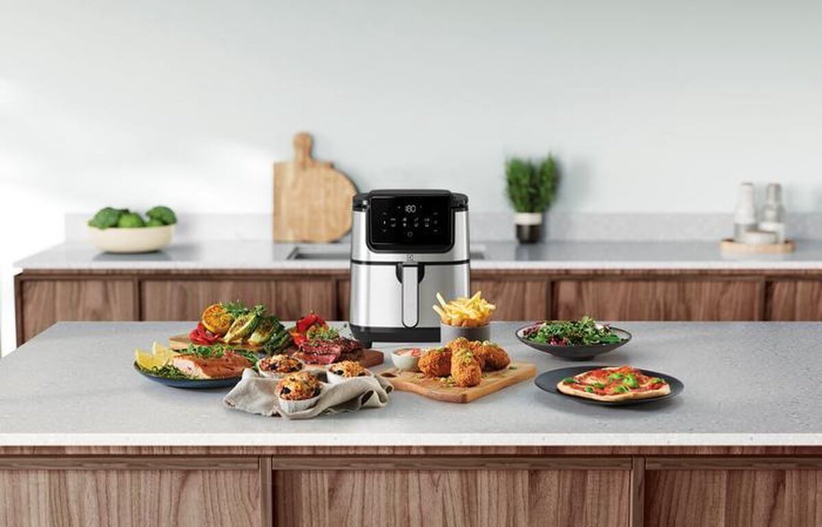 Electrolux E6AF1-4ST airfryer op een keukenblad omringd door gevarieerde gerechten zoals friet, vlees, pizza en salades.
