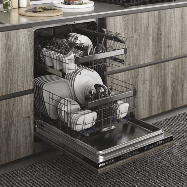 Haier XI 6C3D0FB volledig geïntegreerde vaatwasser met open deur, gevuld met borden, glazen en bestek op drie niveaus, ingebouwd in moderne keuken met houten frontpaneel.