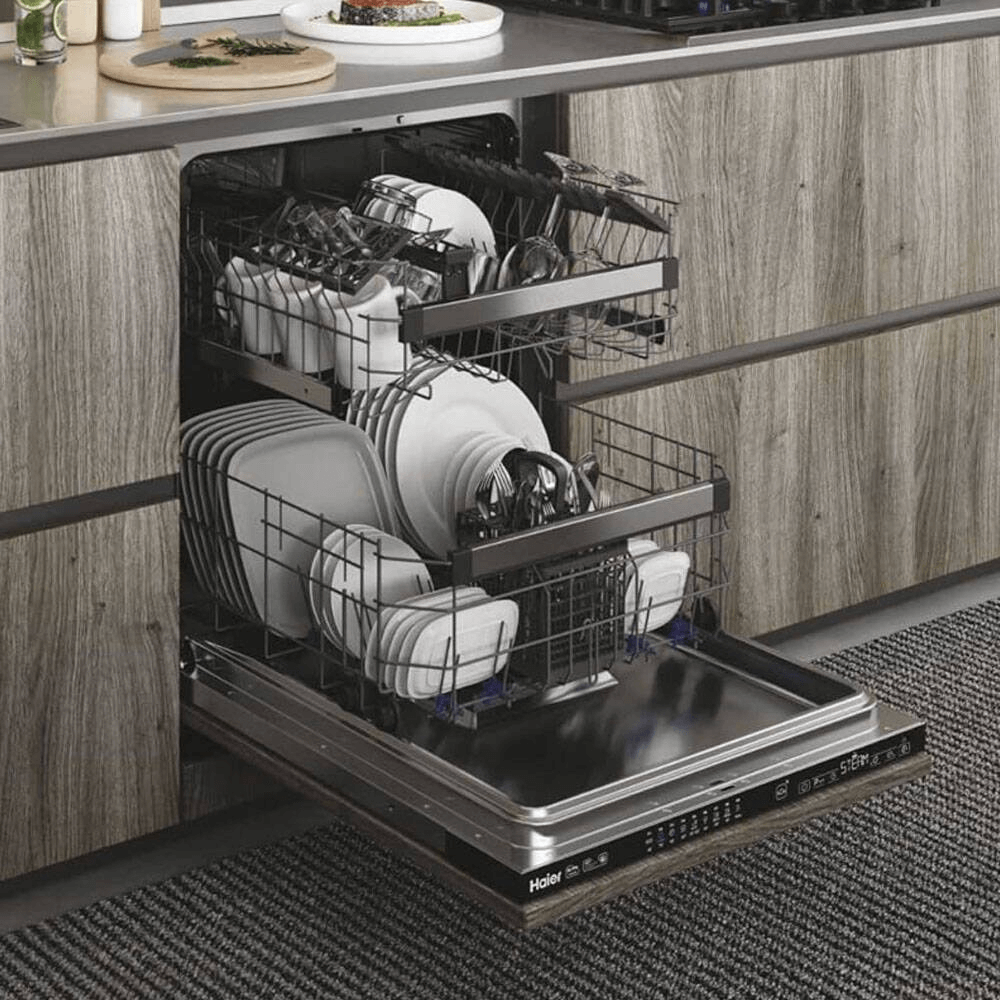 Haier XI 6C3D0FB volledig geïntegreerde vaatwasser met open deur, gevuld met borden, glazen en bestek op drie niveaus, ingebouwd in moderne keuken met houten frontpaneel.