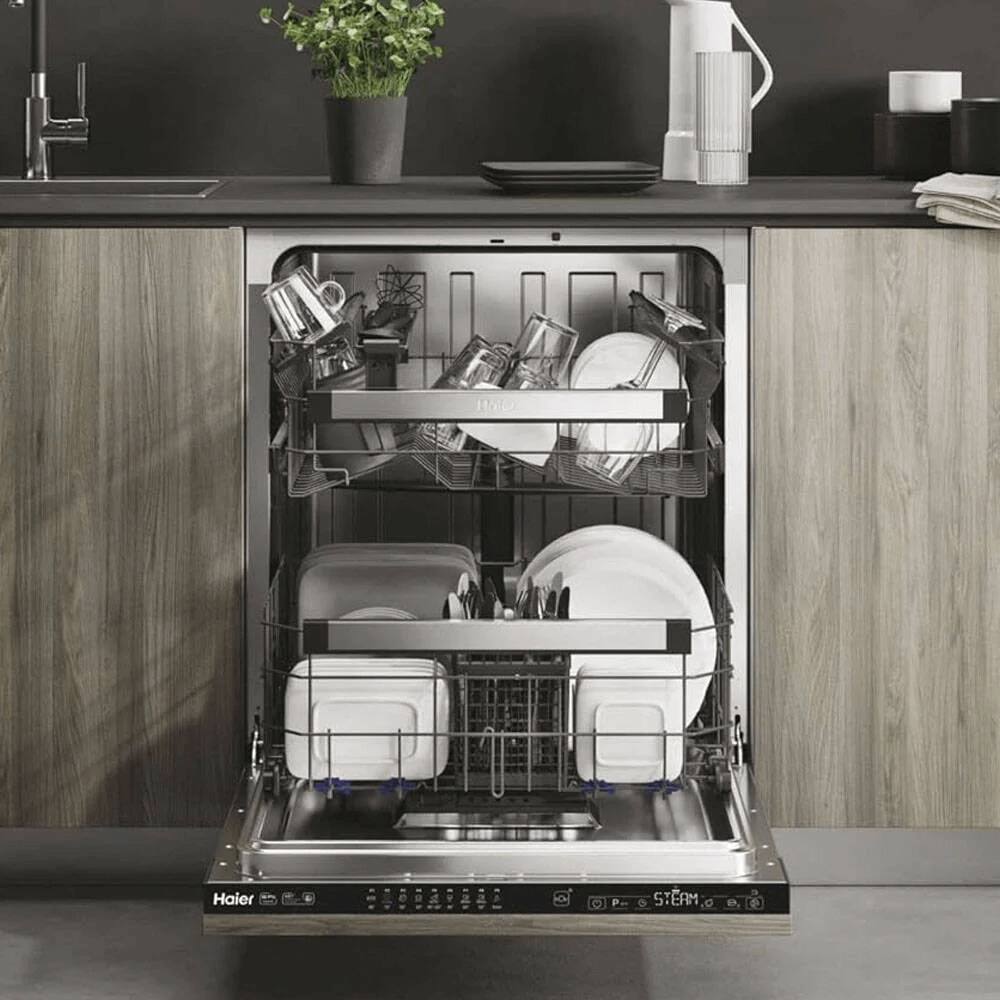 Volledig geïntegreerde Haier XI 6C3D0FB vaatwasser met open deur, gevuld met servies en glazen op drie niveaus, ingebouwd in moderne keuken met houten front en zwarte werkblad.